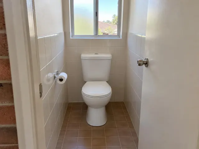Clean white toilet in a small tiled bathroom with window and brown floor tiles in Pagewood