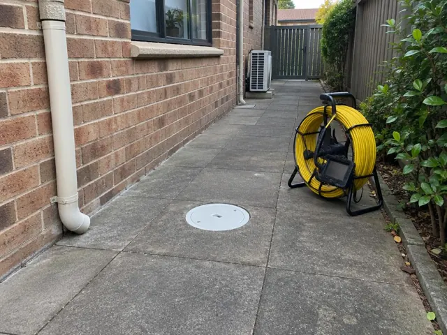 Capped drain inspection opening on a paved side path beside a brick house with a coiled camera reel in Eastgardens