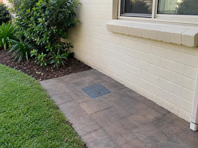 Capped access cover in paved backyard beside a cream brick wall in Pagewood