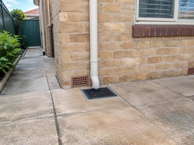 Concrete patio with a seated stormwater grate beneath a white downpipe beside a brick house in Mascot